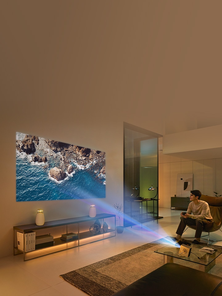Zu sehen ist ein Mann, der auf dem Sofa sitzt und ein 4K-Video genießt, das durch den CineBeam Q HU710PB auf die Wand des Wohnzimmers projiziert wird – zusätzlich ein ein XBOOM Speaker zu sehen.