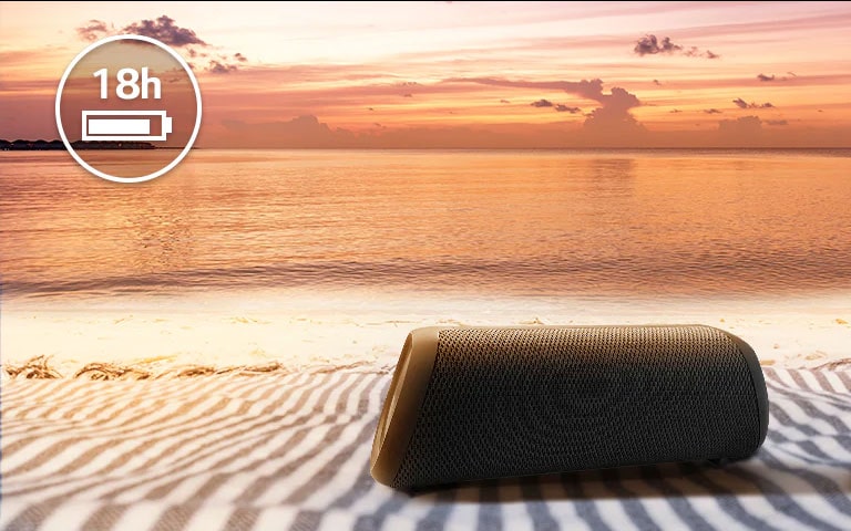 Der Lautsprecher befindet sich auf einem Strandtuch. Im Hintergrund ist ein Sonnenuntergang zu sehen, um zu verdeutlichen, dass das Gerät bis zu 18 Stunden lang betrieben werden kann.