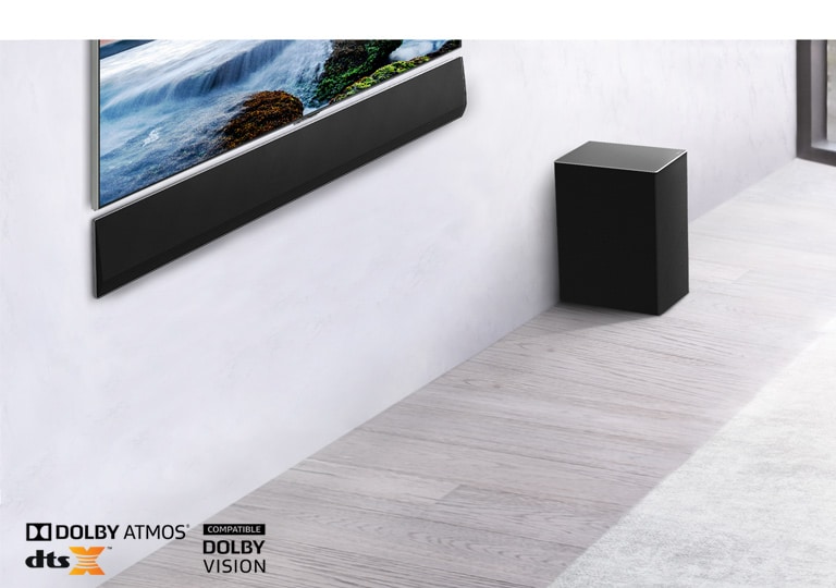 Ein Fernseher ist an der Wand montiert, darunter eine LG Soundbar. Rechts darunter steht ein Subwoofer. Der Fernseher zeigt einen Sonnenuntergang am Meer an.