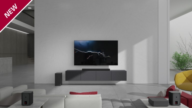 Eine Sound Bar steht in einem Wohnzimmer auf einem grauen TV-Regal unter einem wandmontierten Fernseher. Ein kabelloser Subwoofer steht links davon auf dem Boden und die Sonne scheint von rechts in den Raum. Ein langes Sofa in Rot und Weiß steht gegenüber dem Fernsehgerät und der Sound Bar. In der linken oberen Ecke steht „NEU“.