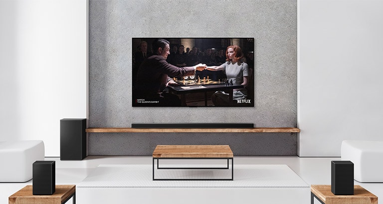 Eine Zusammenstellung, bestehend aus zwei Rücklautsprechern, Subwoofer, einer Soundbar sowie einem TV, befindet sich in einem hellen Wohnzimmer. Ein Poster einer TV-Sendung ist auf dem Bildschirm zu sehen.