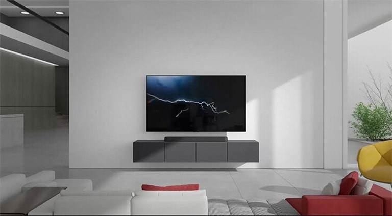 Die Soundbar ist auf dem grauen Schrank in der Mitte platziert und der angeschlossene Fernseher hängt an der weißen Wand direkt darüber. Ein weißes und rotes langes Sofa steht vor dem Fernseher und das Sonnenlicht kommt von der rechten Seite, wo eine grüne Pflanze steht.
