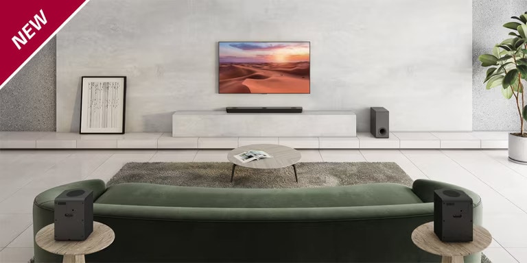 Ein Fernseher hängt an einer weißen Wandverkleidung in einem Wohnzimmer. Darunter steht eine LG Sound Bar auf einem TV-Regal. Auf dem Boden rechts vom Regal steht ein kabelloser Subwoofer. Auf Beistelltischen links und rechts hinter dem Sofa stehen zwei externe Lautsprecher. In der linken oberen Ecke steht „NEU“.