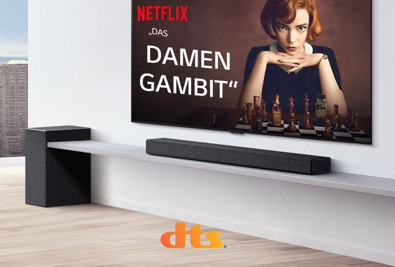 An der Wand befestigter TV.  Ein Poster einer TV-Sendung ist auf dem Bildschirm zu sehen. Die LG Soundbar steht unter dem TV auf einem weißen Regal mit Subwoofer direkt daneben. Die Logos von Dolby Atmos und DTS:X werden in der Mitte unten im Bild angezeigt.