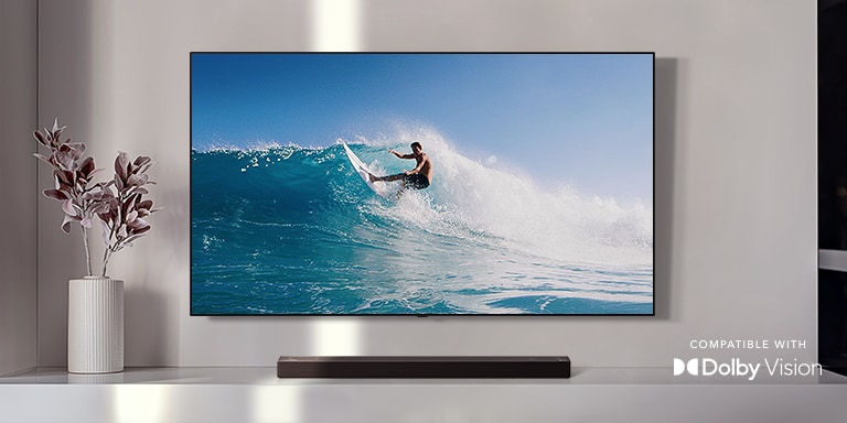 An der Wand befestigter TV. Der TV zeigt einen Mann, der auf einer großen Welle surft. Die LG Soundbar steht direkt unter dem TV auf einem weißen Regal. Direkt neben der Soundbar befindet sich eine Vase mit einer Blume (Video abspielen).