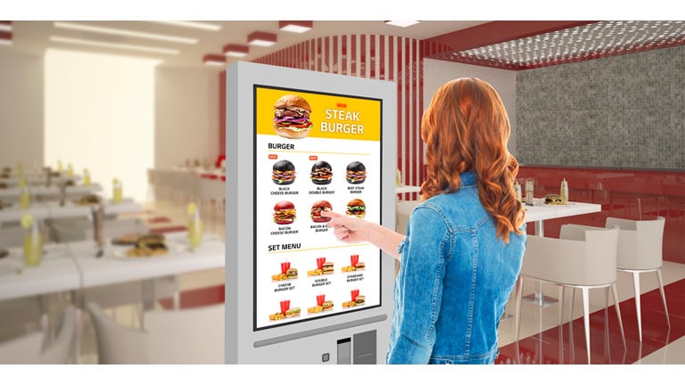 A woman is touching a kiosk to select a hamburger from the menu.
