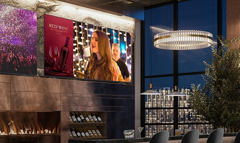 In einer luxuriösen Weinbar sind zwei Displays installiert. Eines zeigt eine Konzertszene, und das andere zeigt zwei Bilder, und zwar sowohl eine Werbung für Rotwein als auch eine Sängerin.