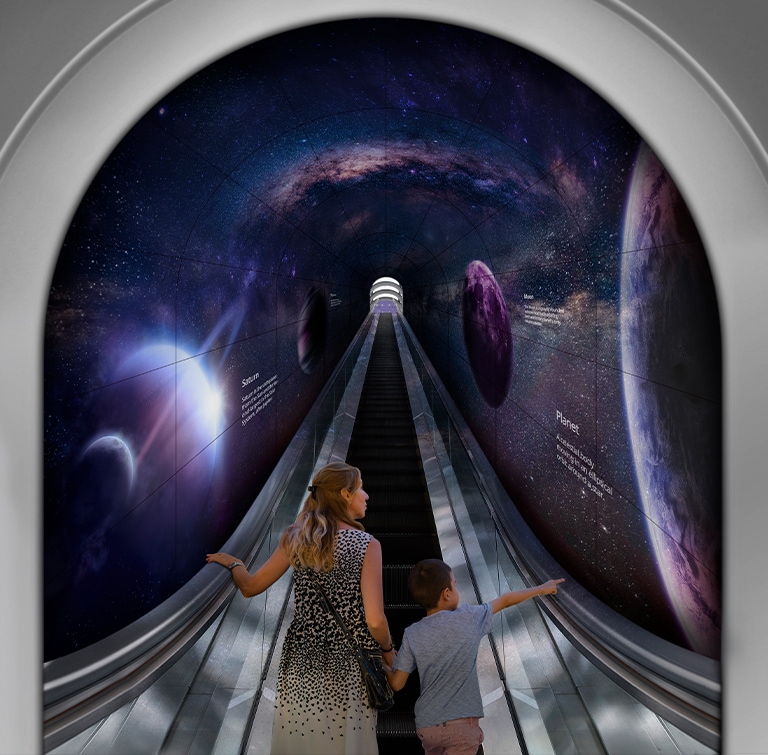 Eine Frau und ein Kind stehen auf einer Rolltreppe, die durch einen Tunnel aus flexibel eingesetzten gebogenen OLED-Elementen im Open-Frame-Stil führt.