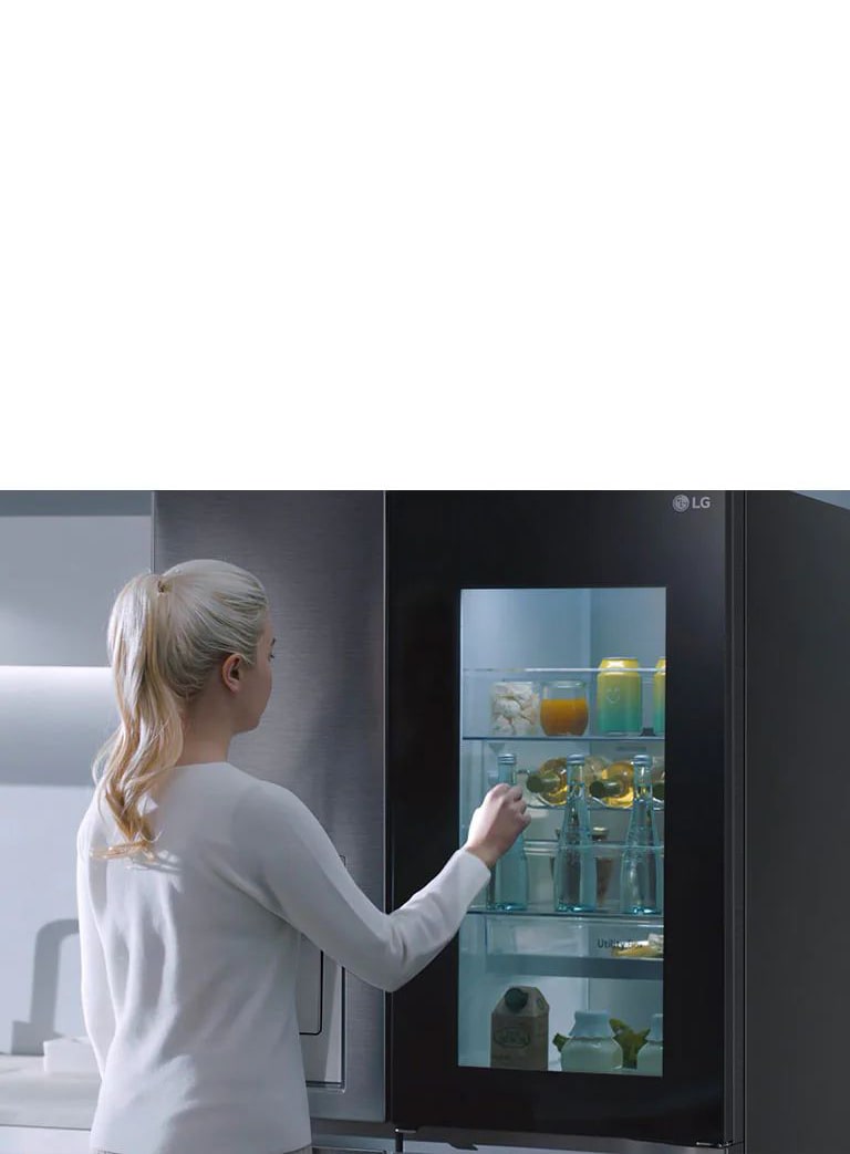 Ein Video zeigt eine Frau, die sich ihrem InstaView-Kühlschrank nähert und zweimal anklopft. Der Innenraum leuchtet auf, und sie kann den Inhalt ihres Kühlschranks sehen, ohne die Tür öffnen zu müssen. Die Ansicht zoomt heran, sodass die Getränke in der Tür zu sehen sind, und zoomt dann heraus, um die Frau von hinten zu zeigen, wie sie die Tür öffnet und sich ein Getränk nimmt.