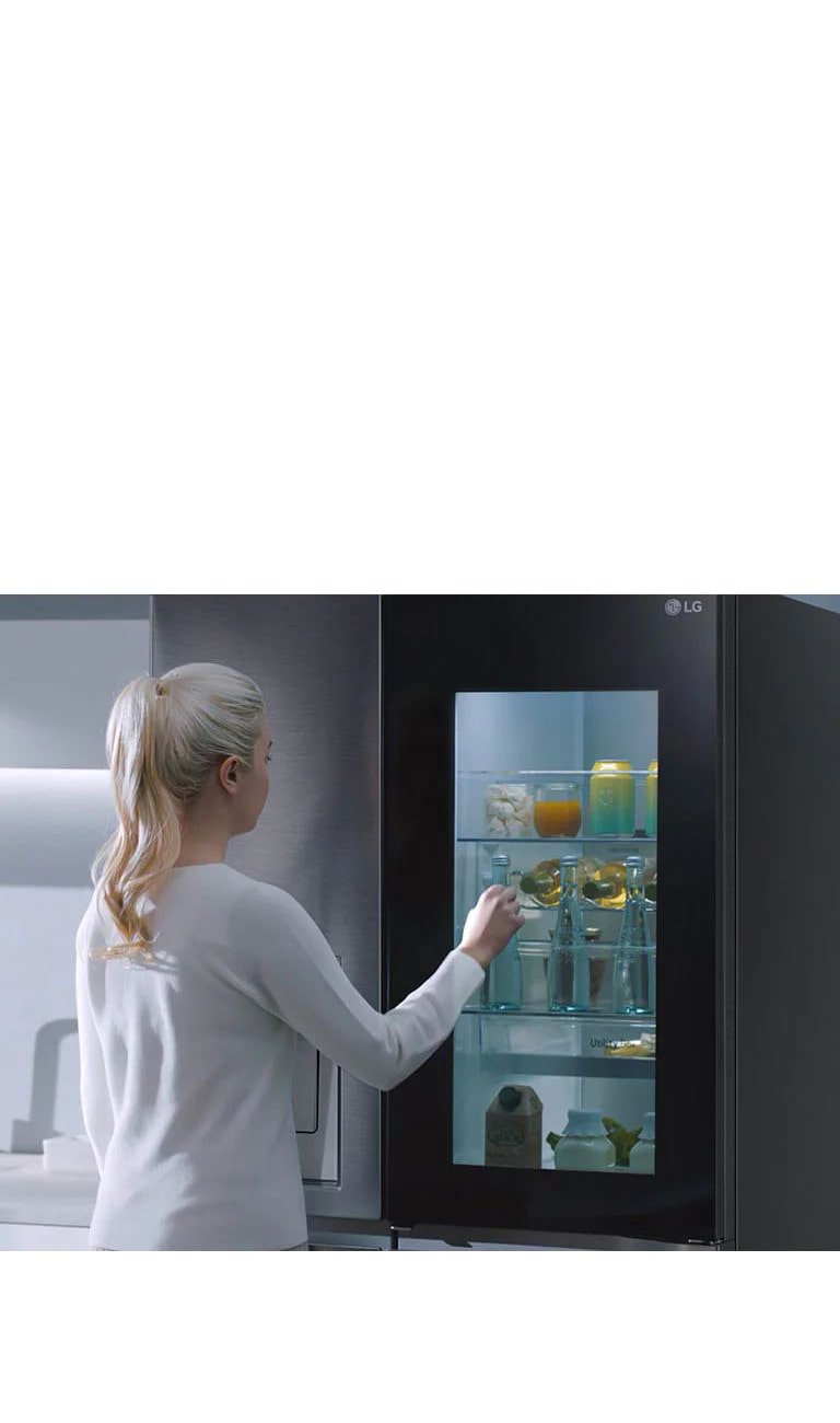 Ein Video zeigt eine Frau, die sich ihrem InstaView-Kühlschrank nähert und zweimal anklopft. Der Innenraum leuchtet auf, und sie kann den Inhalt ihres Kühlschranks sehen, ohne die Tür öffnen zu müssen. Die Ansicht zoomt heran, sodass die Getränke in der Tür zu sehen sind, und zoomt dann heraus, um die Frau von hinten zu zeigen, wie sie die Tür öffnet und sich ein Getränk nimmt.