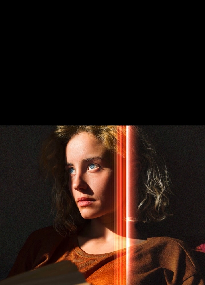Eine Frau mit strahlend blauen Augen und einem orangefarbenen Oberteil in einem dunklen Raum. Rote Linien, die AI-Verfeinerungen darstellen, bedecken einen Teil ihres Gesichts, das hell und detailliert ist, während der Rest des Bildes stumpf aussieht. 	