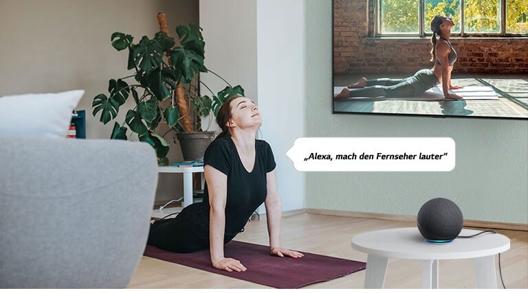 Eine Frau, die im Wohnzimmer vor dem Fernseher Yoga praktiziert, befiehlt einem Smart Speaker, die Lautstärke des Fernsehers zu erhöhen.