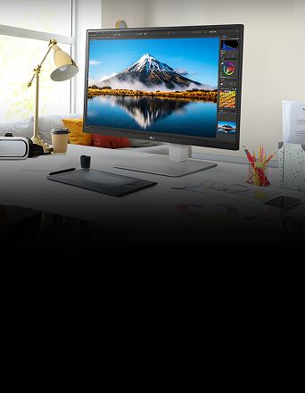 Auf dem Schreibtisch steht ein ultrafeiner Monitor, auf dem Photoshop-Arbeiten zu sehen sind. Neben dem Monitor befinden sich eine Schreibtischlampe, eine Maus, Papier und Buntstifte.