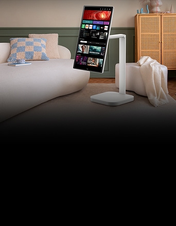 Ein LG Smart Monitor ist im Hochformat in einem Wohnzimmer platziert und zeigt verschiedene Content-Apps an. Der Monitor ist auf einem Schwenkständer montiert, der von einem gemütlichen Innenraum mit Sofa, Kissen und einem Schrank umgeben ist.