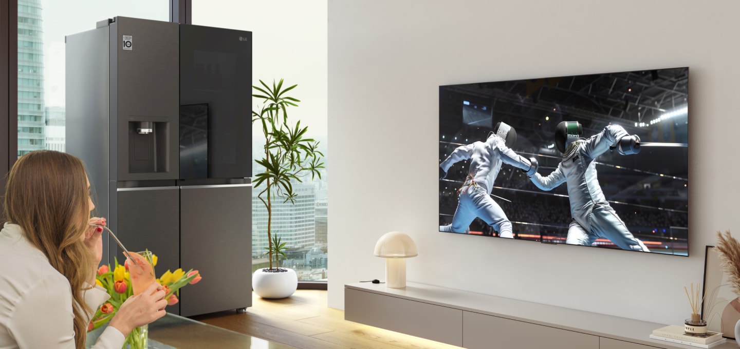 Eine Frau, die sich ein Sportspiel auf einem LG OLED-Fernseher im Wohnzimmer ansieht, mit einem Mocktail in der Hand, mit einem LG InstaView-Kühlschrank neben der Fernsehwand.