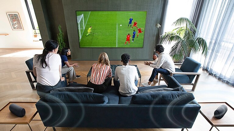5 Personen, die vor einem wandmontierten Flachbildfernseher sitzen und sich ein Fußballspiel ansehen.