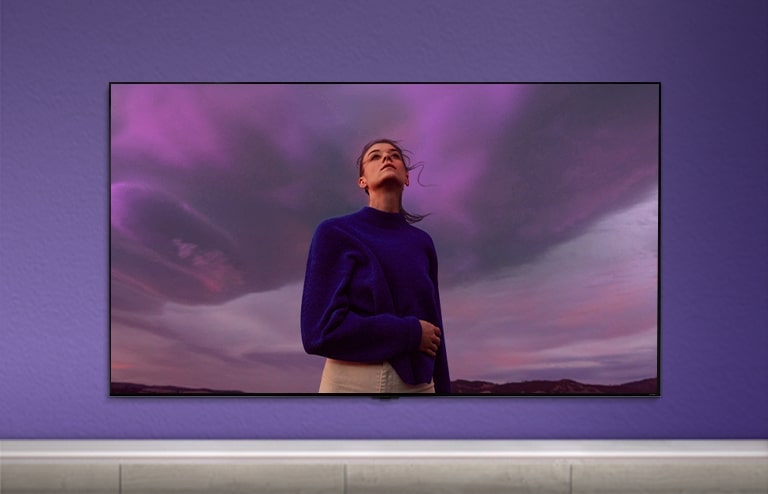 QNED TV ist an einer violetten Wand angebracht und auf dem Bildschirm ist eine Frau in einem violetten Hemd zu sehen. 