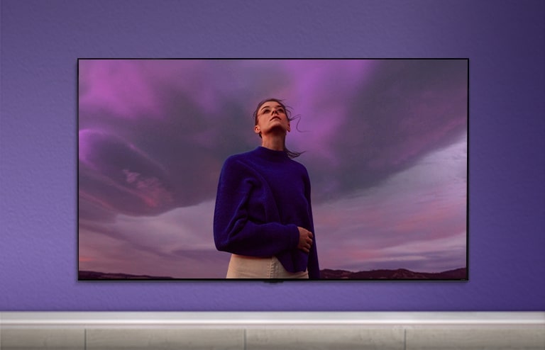 QNED TV ist an einer violetten Wand angebracht und auf dem Bildschirm ist eine Frau in einem violetten Hemd zu sehen.