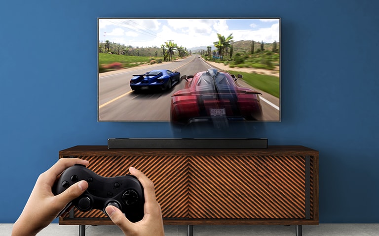 Et LG TV hænger på væggen og viser et racerspil. En LG Soundbar er placeret på den brune hylde lige under LG TV’et. En mand holder et joystick.