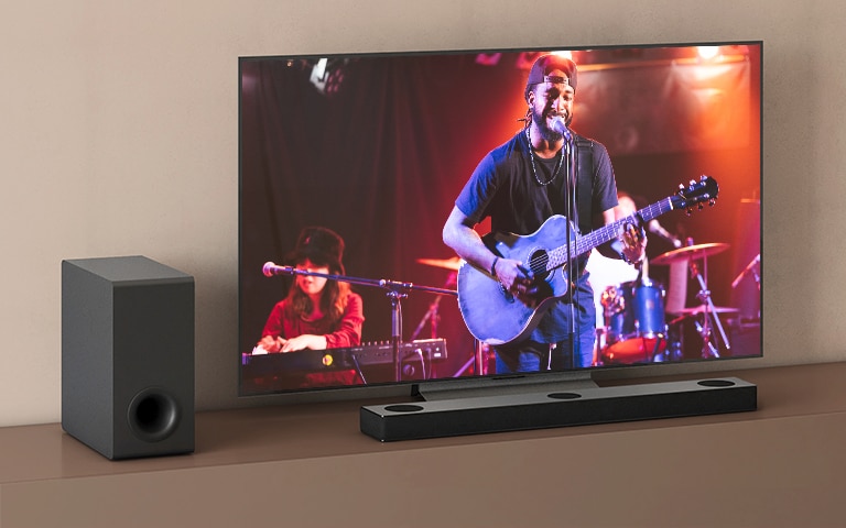 Et LG TV viser en koncert, og en LG Soundbar er placeret under LG TV’et. Til venstre står subwooferen på den brune hylde.