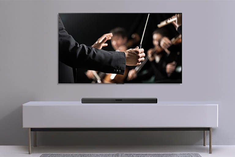 Der vises et TV på en grå væg og med en LG soundbar nedenunder på en grå hylde. TV, der viser en dirigent, som dirigerer et orkester.