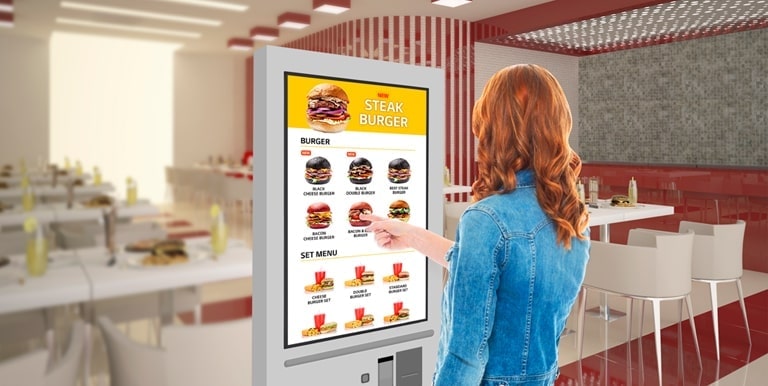 A woman is touching a kiosk to select a hamburger from the menu.