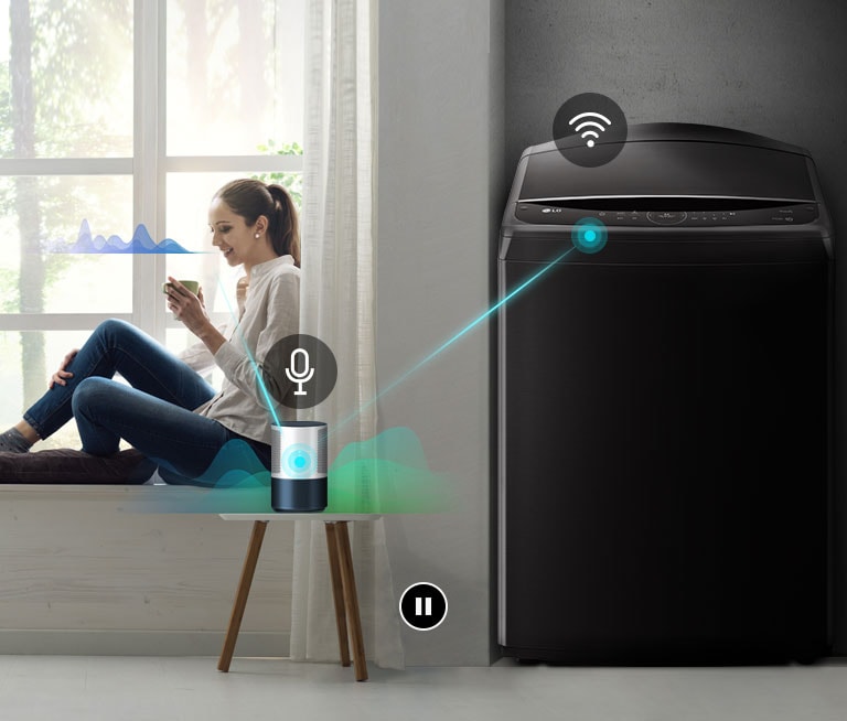 The woman is sitting by the window telling the ai speaker what the washing machine needs.