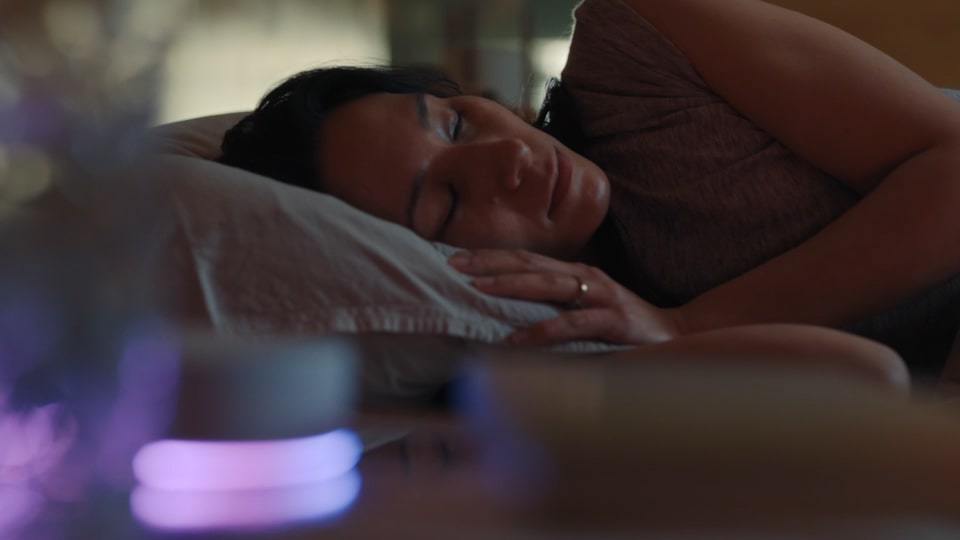 A woman lies on her side, sleeping in bed. In front of her, the LG ThinQ ON device is glowing. 