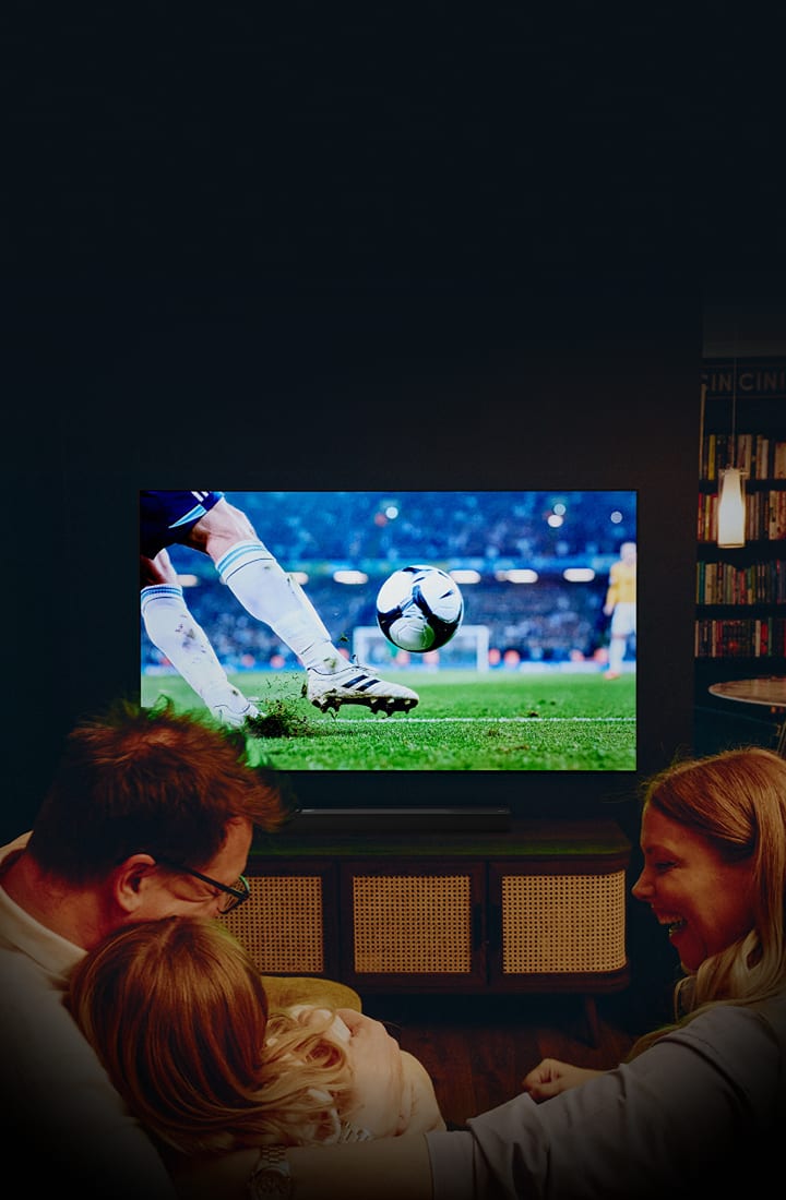 Una familia en un acogedor salón ve un partido de fútbol en un televisor de gran tamaño montado en la pared, mostrando un primer plano de un jugador pateando un balón en el campo de un estadio, con iluminación cálida y una atmósfera cinematográfica.