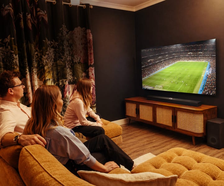 Una familia de tres personas se sienta junta en un sofá en un salón acogedor y tenuemente iluminado, viendo un partido de fútbol en un gran televisor LG OLED. La habitación cuenta con paredes oscuras, cortinas florales, una consola multimedia de madera y altavoces, creando una atmósfera cálida e íntima.