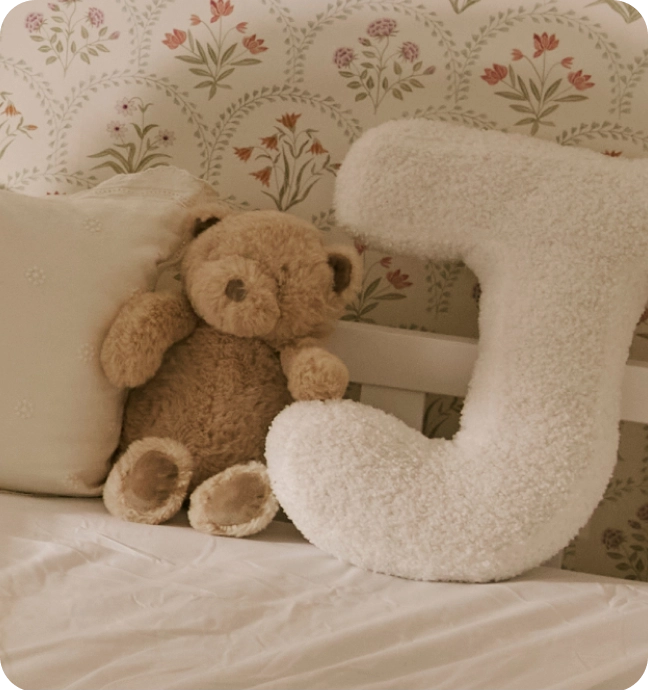 Un osito de peluche marrón y un gran cojín blanco y mullido con la letra 'J' sobre una cama, frente a un papel pintado de flores.