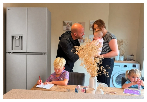 Una familia se reúne en la cocina cerca de un refrigerador de acero inoxidable: un niño dibuja a la mesa, una niña pinta en el mostrador, y los padres están juntos sosteniendo a un bebé.