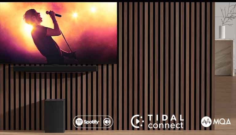 El TV LG OLED C colocado en la pared, con una barra de sonido SC9S debajo colocada en su soporte exclusivo. El subwoofer está debajo. El televisor muestra un concierto.