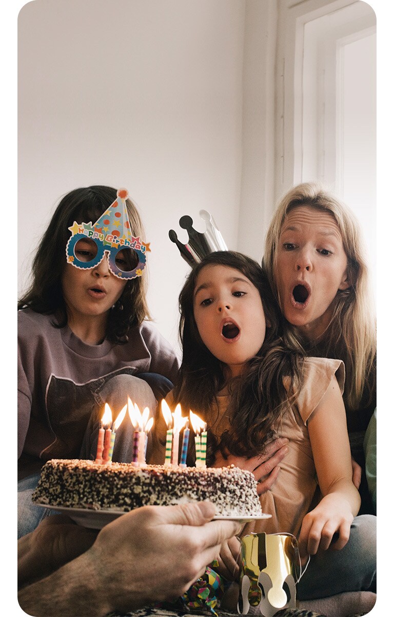 Image of two adult women and a young girl wearing a birthday hat on their head and blowing out the candles on the cake.