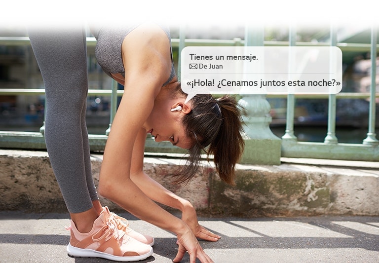 Una mujer está haciendo estiramientos para tocar el suelo mientras escucha un mensaje que los auriculares leen por ella.