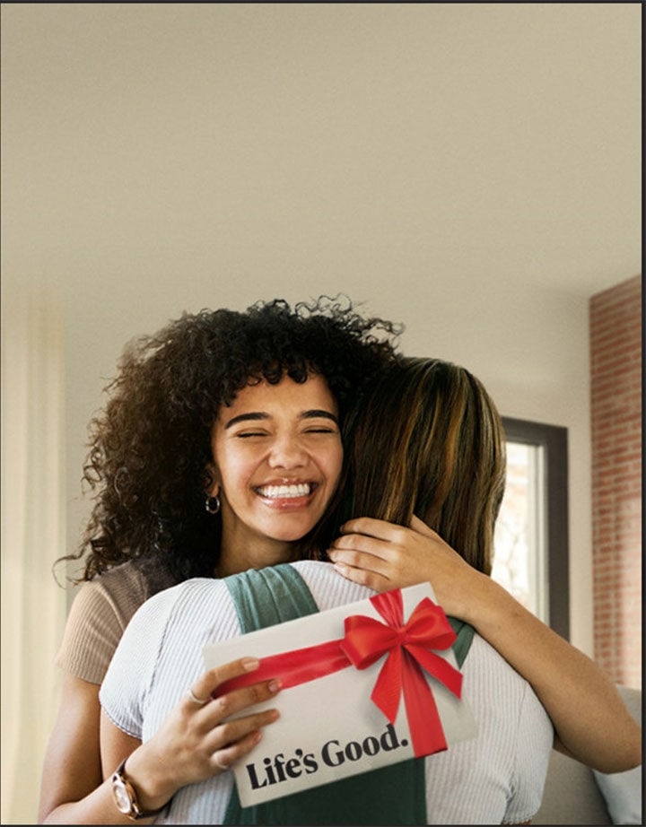 Una mujer sostiene una caja de regalo mientras abraza a una amiga y expresa gratitud. Se la ve agradecida y feliz, y la escena transmite calidez y una fuerte sensación de amistad.	