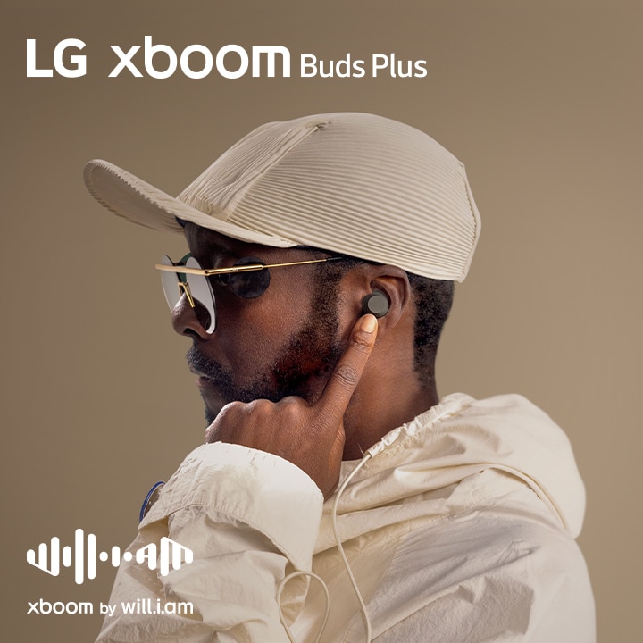 will.i.am en traje blanco y gafas de sol enseñando su lado derecho mirando el auricular en la oreja con el dedo índice izquierdo.