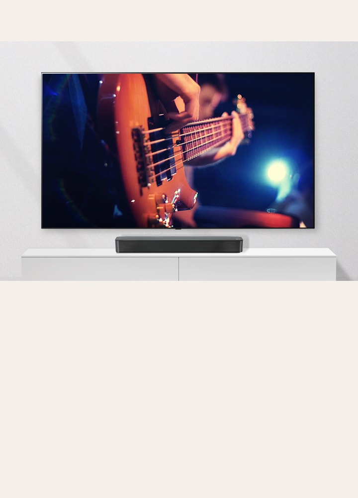 El televisor está pegado a la pared y la barra de sonido está en una estantería blanca. TV que muestra a un hombre tocando la guitarra.