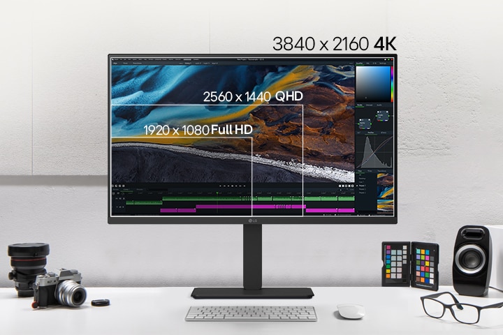 Vista frontal de un monitor LG negro sobre un escritorio, mostrando una comparativa de diferentes resoluciones de pantalla en capas, con cámaras, libros, muestras de color y altavoces dispuestos alrededor.