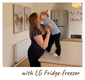 Une mère tient son bébé dans une cuisine moderne, debout à côté d’un réfrigérateur ouvert. [with LG Fridge-freezer]