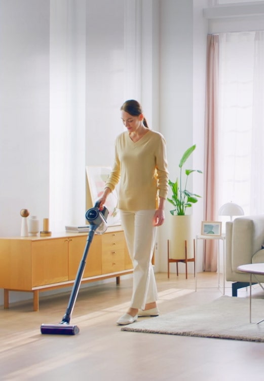 A woman is standing in a living room with both hard floor and carpet and vacuums the floor.