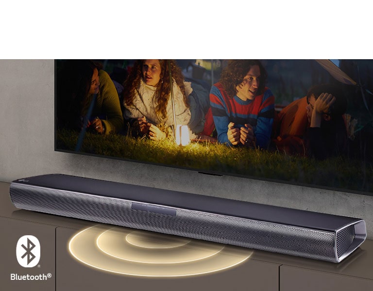 Le téléviseur LG est accroché au mur ; l’écran montre 2 couples allongés sur l’herbe. Devant eux se trouve une lampe. La barre de son LG se trouve sous le téléviseur LG. Un graphique représentant du son sort de l’avant de la barre de son. Le logo Bluetooth est visible dans le coin inférieur gauche de l’image.