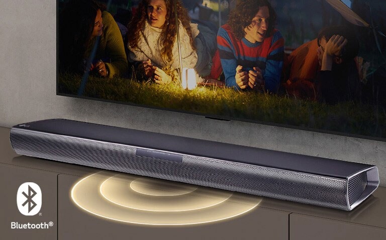 Le téléviseur LG est accroché au mur ; l’écran montre 2 couples allongés sur l’herbe. Devant eux se trouve une lampe. La barre de son LG se trouve sous le téléviseur LG. Un graphique représentant du son sort de l’avant de la barre de son. Le logo Bluetooth est visible dans le coin inférieur gauche de l’image.