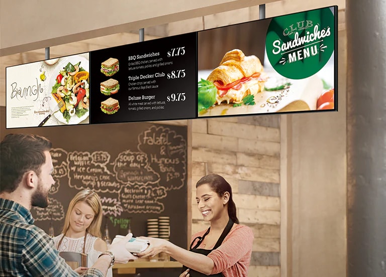 Le personnel d’un magasin de sandwichs remet un sandwich à un client. Au-dessus d’eux, la série SM5J indiquant un panneau de menu a été installée et affiche des menus avec promotions sur les brunch.