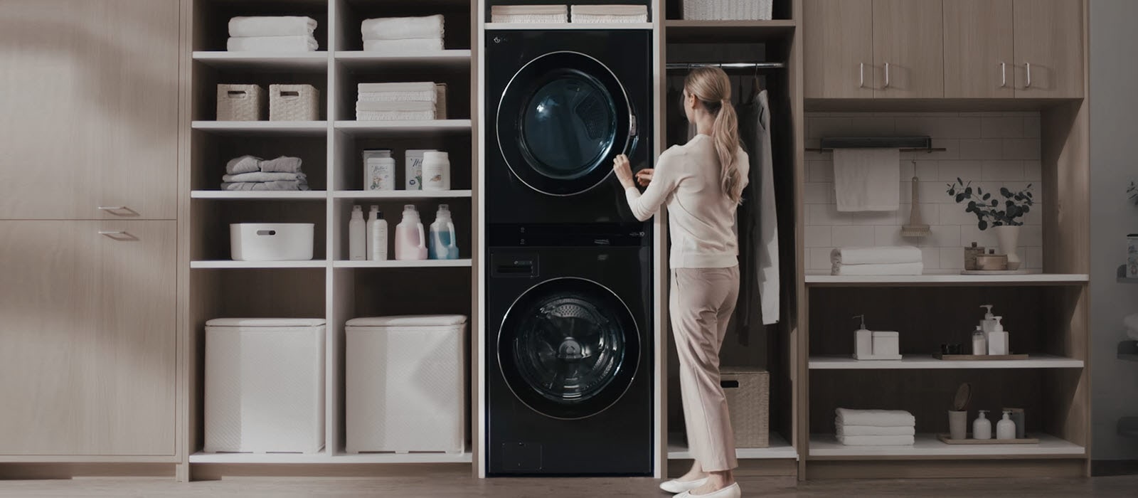 Un lave-linge et un sèche-linge blancs sont empilés l’un sur l’autre et installés dans une unité murale avec des étagères et un placard et une femme a le dos tourné vers l’avant alors qu’elle se lève pour appuyer sur un bouton du panneau de commande supérieur central. Sa position est inconfortable.