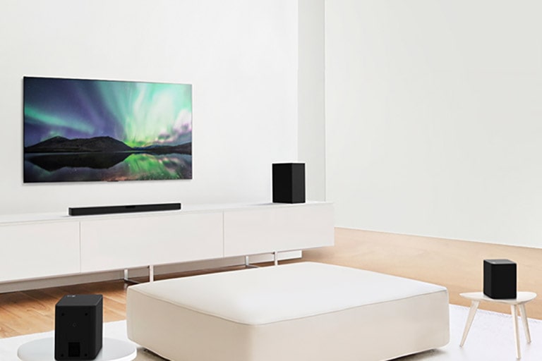 Vue du TV et de la  barre de son dans un salon blanc avec un canapé blanc au centre. Des haut-parleurs sont placés aux deux extrémités du canapé.