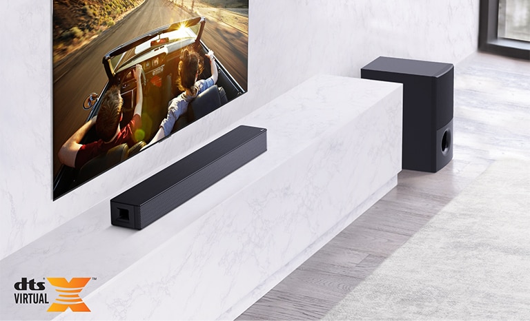 Le téléviseur est sur le mur gris, la barre de son LG est en dessous sur une étagère en marbre blanc avec un caisson de basse à droite. Le téléviseur montre un couple dans une voiture.