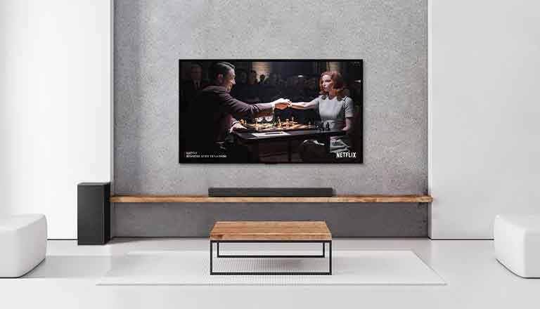 Un caisson de graves, une barre de son et un téléviseur sont dans un salon blanc. Une femme et un homme jouent aux échecs sur l'écran TV.