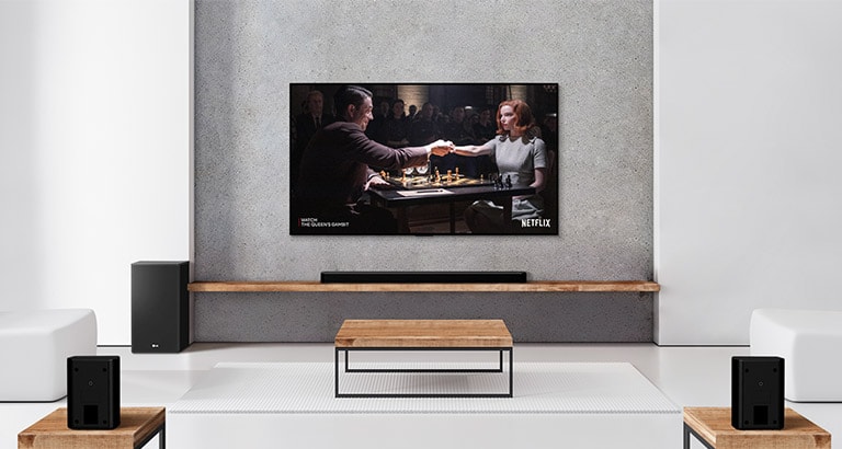 Un ensemble de 2 haut-parleurs arrière, un caisson de basses, une barre de son et un téléviseur se trouvent dans un salon blanc. Une femme et un homme jouent aux échecs sur un écran de télévision.