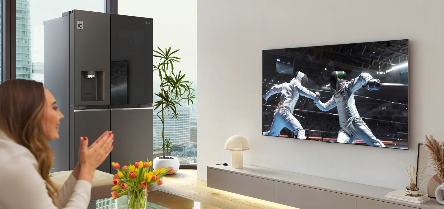Une femme regardant un match de sport sur un téléviseur LG OLED dans le salon, tenant un Boissons sans alcool, avec un réfrigérateur LG InstaView à côté du mur du téléviseur.